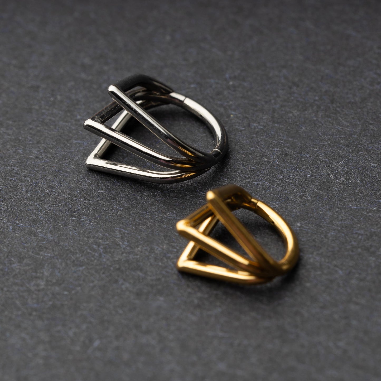 Close-up of two geometric septum clickers in silver and gold, featuring a unique layered triangular design, displayed on a dark background.
