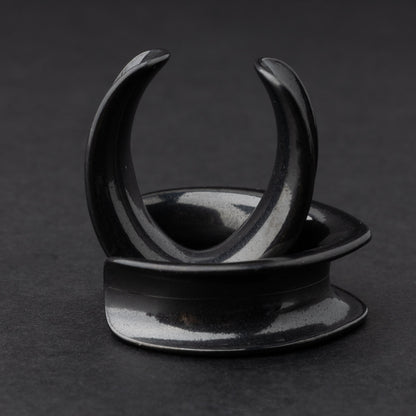 A collection of double-flared metal tunnels for stretched ears in black finish, displayed on a dark background.
