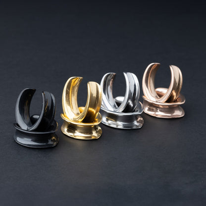 A collection of double-flared metal tunnels for stretched ears in gold, silver, black and rose gold  finish, displayed on a dark background.
