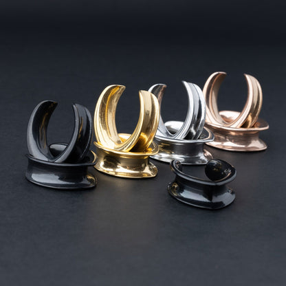 A collection of double-flared metal tunnels for stretched ears in gold, silver, black and rose gold  finish, displayed on a dark background.
