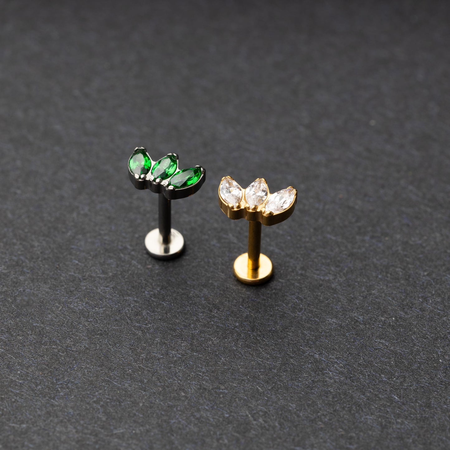 Two flat back bars featuring a triple gem design: one with emerald green gems on a silver base and the other with clear gems on a gold base, displayed on a dark background.
