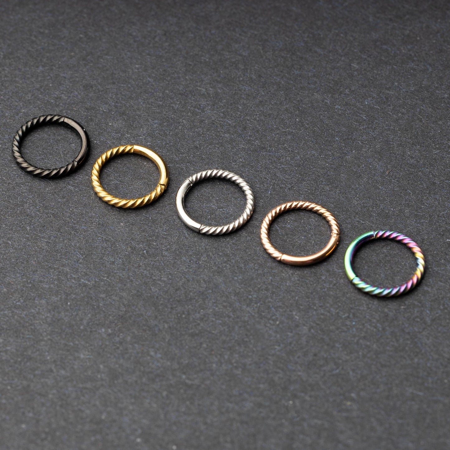 Twisted seamless hoop rings in black, gold, silver, rose gold, and rainbow finishes displayed on a dark background, highlighting their elegant and textured design.