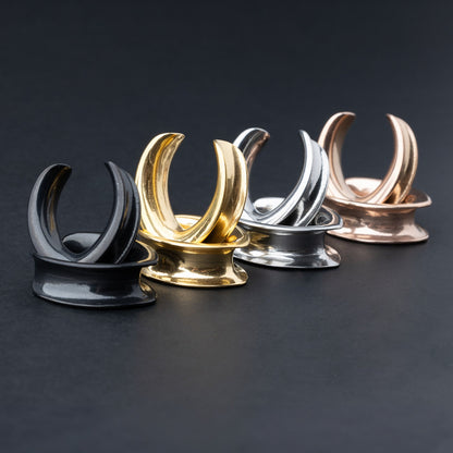 A collection of double-flared metal tunnels for stretched ears in gold, silver, black and rose gold  finish, displayed on a dark background.
