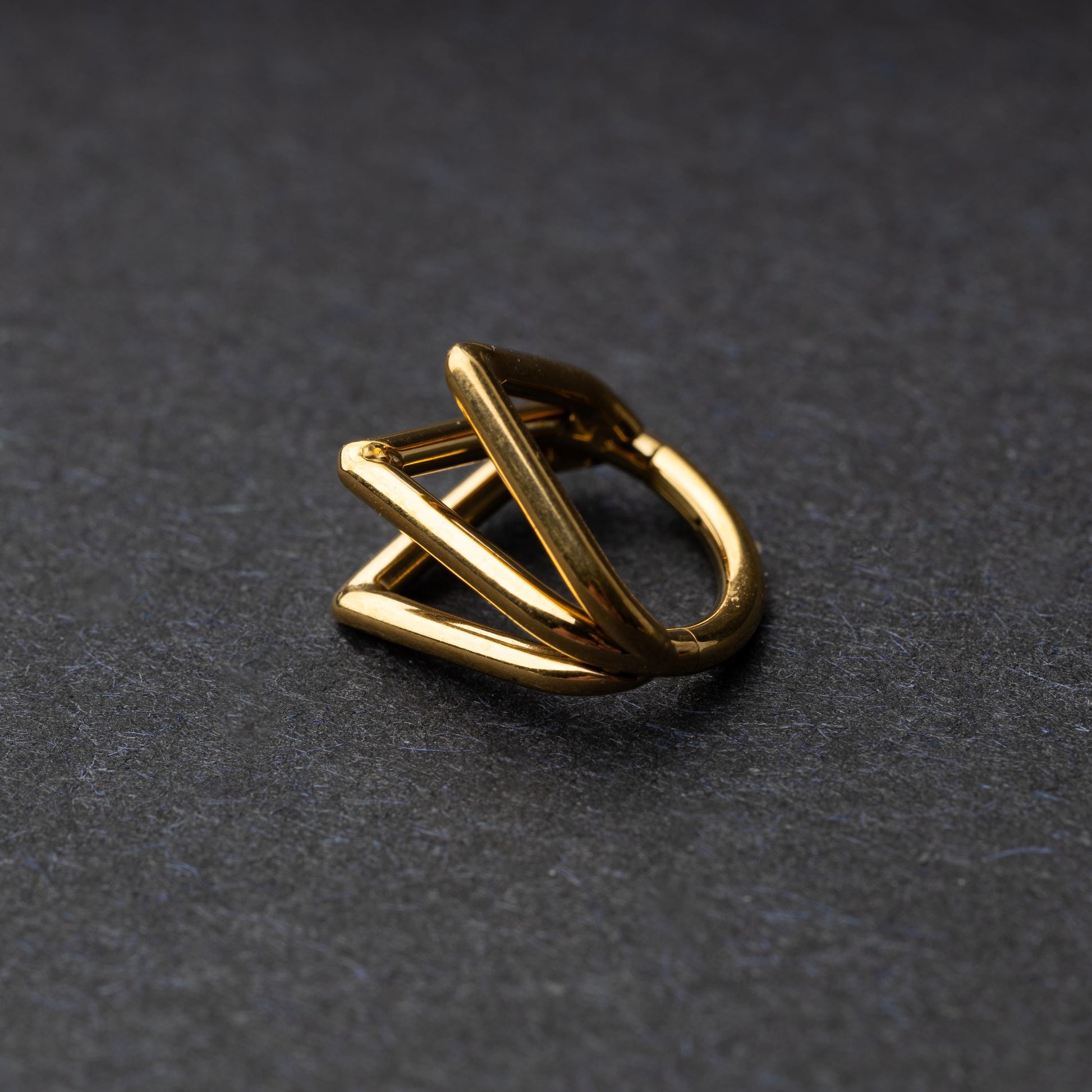 Close-up of a geometric septum clicker in gold, featuring a unique layered triangular design, displayed on a dark background.

